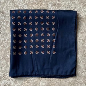 May include: Navy blue pocket square with a repeating pattern of small, copper-colored circles. The square is folded neatly, showcasing the detailed design. This accessory is suitable for formal or business attire.