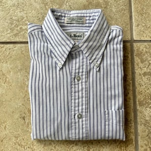 Puede incluir: Una camisa abotonada doblada con rayas verticales azules y blancas. La camisa tiene cuello abotonado, bolsillo en el pecho y la marca "L.Bean" es visible en el cuello.