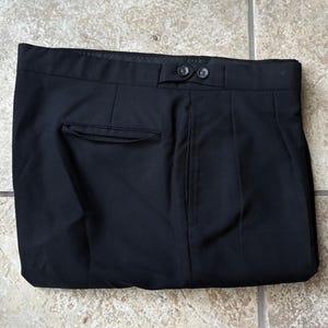 May include: A pair of black trousers featuring a structured waistband with two dark buttons, a single horizontal welt pocket on the right side, and a clean, flat-front design. The fabric appears smooth and solid black throughout the garment.