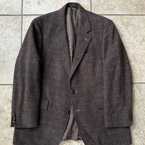 May include: A dark brown blazer featuring a subtle windowpane check pattern in muted red and blue tones. The garment includes notched lapels, two front buttons, two flap pockets, and four small buttons on each sleeve cuff, with a solid brown lining.