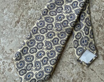 May include: A cream-colored tie with a repeating paisley pattern in navy blue. The tie is made of a smooth fabric and is lying on a neutral-toned surface. The tie is a classic accessory.