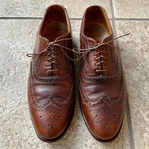 Puede incluir: Un par de zapatos de vestir de cuero marrón con intrincadas perforaciones y cierres con cordones. Los zapatos tienen un diseño clásico y un acabado pulido, adecuados para ocasiones formales.