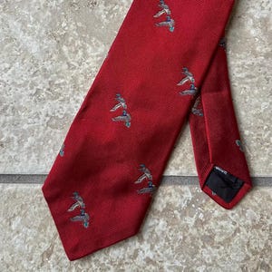 May include: A red necktie with a repeating pattern of small, stylized figures. The tie is made of a textured fabric and is angled across a neutral-toned surface. The figures are in shades of gray and blue.
