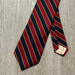 May include: A necktie featuring a diagonal striped pattern in dark navy blue and deep red, accented by thin yellow lines. The tie is constructed from a textured fabric and includes a visible white label on the interior of the narrow end.