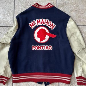 Pode incluir: Uma jaqueta de beisebol azul marinho com mangas de couro branco. A jaqueta tem listras vermelhas e brancas ao redor da bainha e dos punhos. A parte de trás da jaqueta tem um remendo bordado vermelho com o texto "McMahon Pontiac" e uma cabeça de nativo americano.