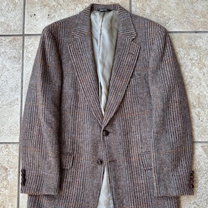 May include: A brown tweed blazer with a subtle plaid pattern. The jacket features a notched lapel, two front pockets, and a two-button closure. The lining is a light green color. The sleeves have three buttons at the cuff.