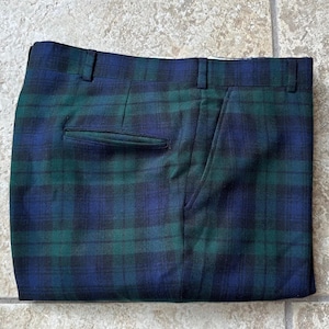 Puede incluir: Pantalones a cuadros verdes y azules doblados. Los pantalones presentan un diseño clásico de tartán. Tienen un corte clásico con cierre de botón y cremallera, trabillas y bolsillos. La tela tiene una textura tejida.