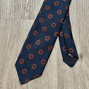 Puede incluir: Una corbata azul marino con un patrón repetido de círculos color óxido. La corbata está hecha de una tela suave y está en ángulo sobre una superficie de madera. Un accesorio clásico y versátil.