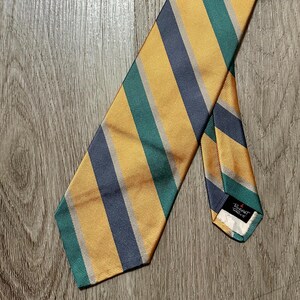 Peut inclure: Une cravate rayée avec des bandes alternées de jaune, de vert d'eau et de bleu marine. La cravate est posée sur une surface en bois, mettant en valeur son motif. L'extrémité de la cravate est visible, avec une petite étiquette.