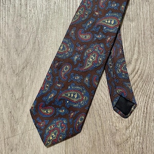 May include: A brown paisley tie featuring a repeating pattern of blue, red, and yellow floral designs. The tie is made of a smooth fabric and is displayed on a wooden surface.