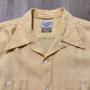 May include: A light yellow linen button-down shirt with two chest pockets and a wide collar. The shirt has a textured appearance and features a label with text. The shirt is made of linen.