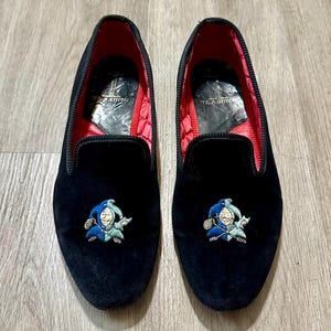 May include: A pair of black velvet loafers with a red quilted lining. Each shoe features an embroidered jester design in blue, white, and green. The soles are black, and the shoes are displayed on a wooden surface.