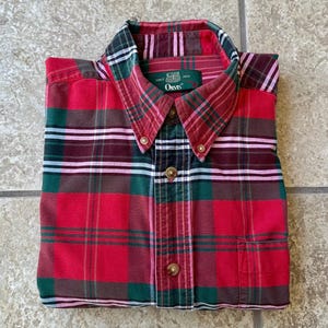 May include: A folded red, green, and brown plaid button-down shirt. The shirt has a button-down collar and a chest pocket. The brand name "Orvis" is visible on a tag near the collar.