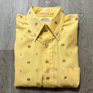May include: A bright yellow button-down shirt featuring a repeating brown pattern of small animal faces and circular icons. The shirt has a pointed collar, a front button placket, and a single chest pocket. The inner label reads Permanent Press.