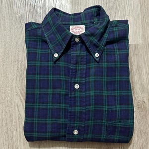 Könnte beinhalten: Ein langärmeliges Hemd mit Knopfleiste und einem dunkelblau-grünen Karomuster. Das Hemd verfügt über einen Button-Down-Kragen, eine durchgehende Knopfleiste und eine Brusttasche. Ein weißes Etikett am Kragen zeigt Brooks Brothers 15-R.