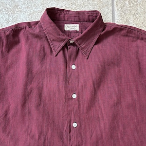 May include: A burgundy button-down shirt with vertical stripes. The shirt has a pointed collar and three visible white buttons. The label on the collar reads "Fast colour shirt".