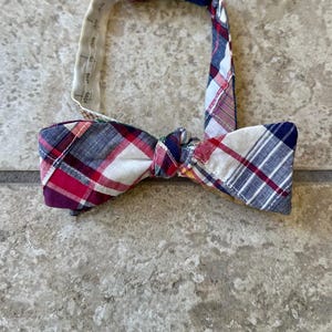 May include: A plaid bow tie in red, white, and blue. The bow tie has a classic shape with a pre-tied knot. The adjustable neckband is white with printed measurements. The bow tie is made of a lightweight fabric.