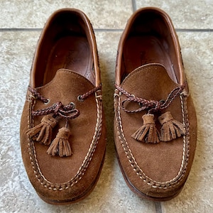 May include: A pair of brown suede loafers with decorative tassels and stitched detailing. The shoes have a classic design, with a leather interior and a rubber sole. The loafers are a size 43.