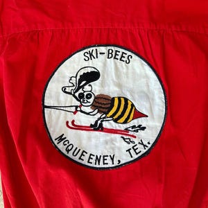 May include: Red short-sleeved shirt with an embroidered patch featuring a bee skiing. The patch reads "SKI-BEES" above the bee and "MCQUEENEY, TEX." below. The bee is brown and yellow, with a black hat and skis.