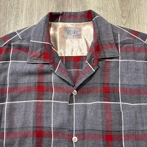 May include: A gray, red, and white plaid button-up shirt. The shirt has a camp collar and a single chest pocket. The label inside the collar reads "Blocks". The shirt is made of a soft, possibly flannel, material.