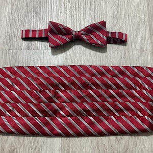 May include: A formal bow tie and a matching cummerbund set featuring a deep red fabric with a diagonal pattern of thin white and dark blue stripes. The bow tie is pre-tied with an adjustable strap, and the cummerbund has horizontal pleats.