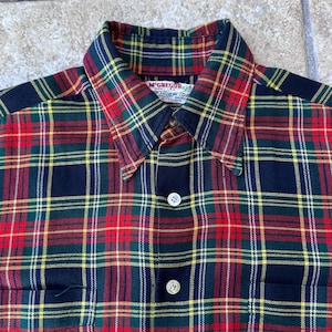 May include: A button-down plaid shirt in red, green, yellow, and navy blue. The shirt features a classic plaid pattern, a collar, and two white buttons. The shirt has a pocket on the left side. The brand name "McGregor" is visible on the collar.