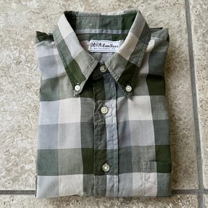 May include: A folded button-down shirt with a green, grey, and white plaid pattern. The shirt has a button-down collar and a chest pocket. The label reads "R.K. Tom Bass, Bethlehem, PA."