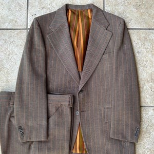 May include: A brown pinstripe suit with a matching jacket and trousers. The jacket features a notched lapel, two buttons, and three buttons on each cuff. The interior lining is striped with orange and green. The trousers have a front pocket and a belt loop.