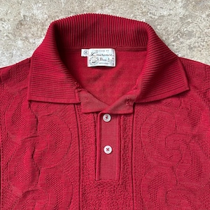 May include: A red collared shirt with a textured pattern and two white buttons. The shirt has a ribbed collar and a label that reads "Laurence B. Jr."