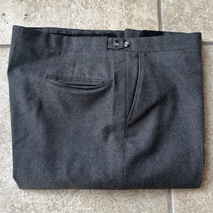 May include: A pair of charcoal gray trousers featuring a flat front, a waistband with a button closure, a single welt pocket on the right side, and a side slash pocket. The fabric has a textured, heathered appearance with a consistent dark gray tone.
