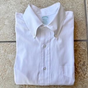 Może przedstawiać: Krystalicznie biała koszula zapinana na guziki z kołnierzykiem zapinanym na guziki i kieszenią na piersi. Koszula jest starannie złożona, podkreślając jej czyste linie i klasyczny design. Etykieta głosi "16-2 Brooks Brothers".