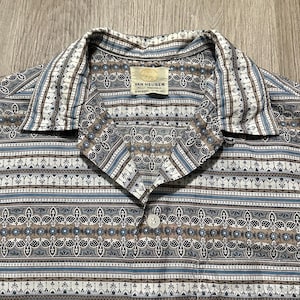 Pode incluir: Close-up de uma camisa vintage com botões e padrão. A camisa tem gola branca e fecho de botão. O padrão inclui formas geométricas azuis, castanhas e brancas. A etiqueta diz "Van Heusen".