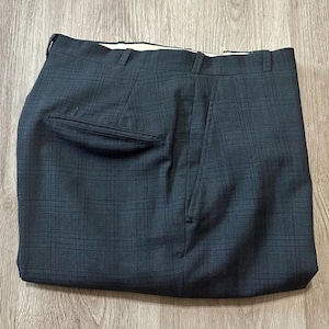 May include: Dark charcoal gray trousers featuring a subtle black windowpane plaid pattern. The pants include a structured waistband with belt loops, a single rear welt pocket with a flap, and a clean, tailored cut with a flat front design.