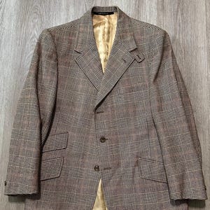 May include: A brown and tan plaid blazer featuring a notched lapel, a three-button front closure, and two flap pockets on the lower front. The jacket is lined with a solid gold-colored fabric and includes a small tab detail on the left lapel.