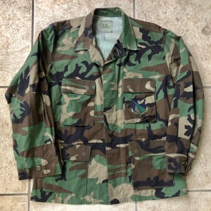 Könnte beinhalten: Eine grüne, braune und schwarze Tarnjacke mit einem Aufnäher auf der linken Tasche. Der Aufnäher ist grün, blau und weiß mit einem geflügelten Symbol und dem Text "335th Tactical Fighter Squadron".