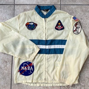 May include: Cream-colored vintage jacket with blue and white stripes and patches. Features a NASA logo, a US flag, and other space-themed patches. The jacket has a zip-up front and a stand-up collar.