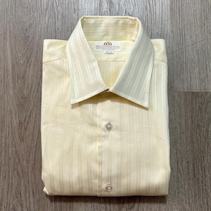 May include: A pale yellow long-sleeve button-down shirt featuring a subtle vertical stripe pattern. The shirt has a pointed collar and a front placket with visible buttons. A white label inside the collar reads The Custom Shop.