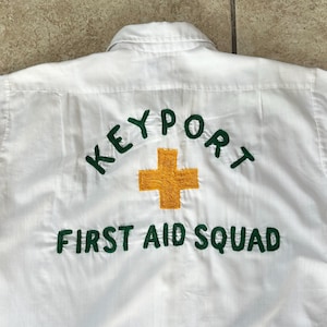 May include: White shirt with the text "KEYPORT FIRST AID SQUAD" embroidered in green. A gold cross is centered between the text. The shirt has a collar and appears to be made of a lightweight fabric.