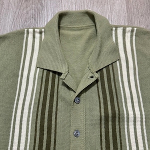 May include: Olive green button-down knit shirt with vertical stripes in white and dark green. The shirt has a collared neckline and two buttons. The knitwear is visible in the close-up shot.