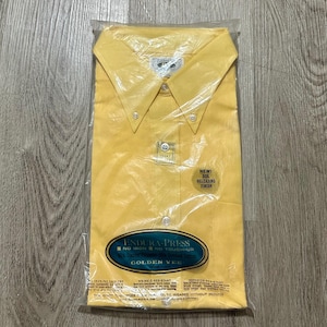 May include: A new, yellow button-down shirt in a clear plastic package. The shirt has a button-down collar and a chest pocket. The packaging includes the text "ENDURA-PRESS" and "GOLDEN VEE". The packaging also states "NEW! SOIL RELEASING FINISH".