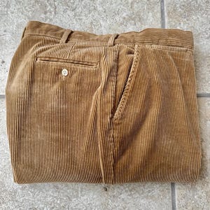 Pode incluir: Calças de veludo cotelê marrons dobradas com um bolso traseiro abotoado e bolsos laterais. As calças têm um design clássico e um tecido texturizado e canelado. A cor é um tom quente e terroso.