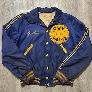 May include: Vintage blue jacket with yellow accents, featuring a basketball emblem and the text "CWV 1952-53." The jacket has the name "Jackie" embroidered in script. It has yellow stripes on the sleeves and around the cuffs and waist.