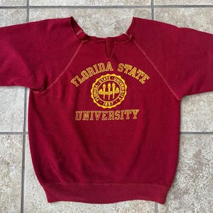 May include: A maroon short-sleeved sweatshirt featuring a yellow and gold emblem that reads "FLORIDA STATE UNIVERSITY 1857". The sweatshirt has a crew neck with a small v-shaped cutout.