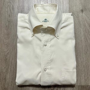 Könnte beinhalten: Ein cremefarbenes Hemd mit Button-Down-Kragen, langen Ärmeln und einer einzelnen Brusttasche. Das Etikett am Innenkragen trägt den Text Borrelli Napoli. Das Hemd ist mit einer Knopfleiste und hellen Knöpfen ausgestattet.