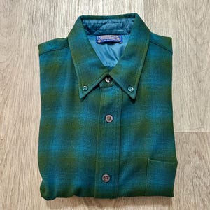 May include: A long-sleeve button-down shirt featuring a teal and olive green plaid pattern. The shirt has a pointed collar with button-down tips, a single chest pocket, and a quilted teal interior lining. A Pendleton brand label is visible at the neck.