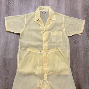 May include: A two-piece yellow pajama set. The top is a short-sleeved button-down shirt with a chest pocket. The shorts have an elastic waistband and button closure. The fabric has a textured, semi-sheer appearance.