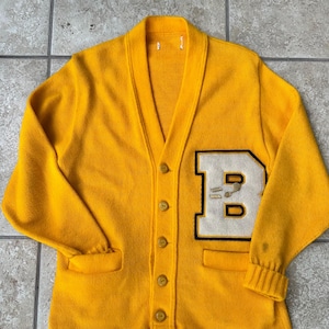 Puede incluir: Un cárdigan amarillo vintage con cuello en V y cierre de botones. El suéter presenta un gran parche blanco y negro con la letra "B" en el pecho izquierdo. Los puños y el ribete del bolsillo son acanalados.