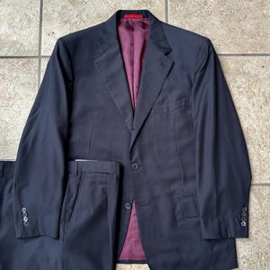 May include: A navy blue pinstripe suit, including a blazer and trousers, is displayed. The blazer has a two-button closure and a burgundy lining. The suit is laid out on a light-colored tiled surface.