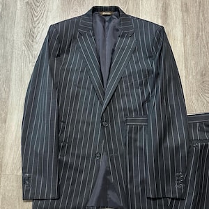 May include: A dark charcoal gray suit featuring a white pinstripe pattern. The jacket includes a notched lapel, two front buttons, and three buttons on each sleeve cuff. The matching trousers are visible on the right side of the jacket.