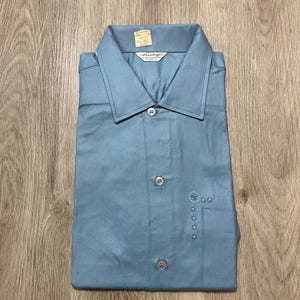 May include: A light blue button-down shirt with a pointed collar and a front placket featuring three visible buttons. The shirt includes a small embroidered floral design on the left chest area and a brand label inside the collar reading Pearly.
